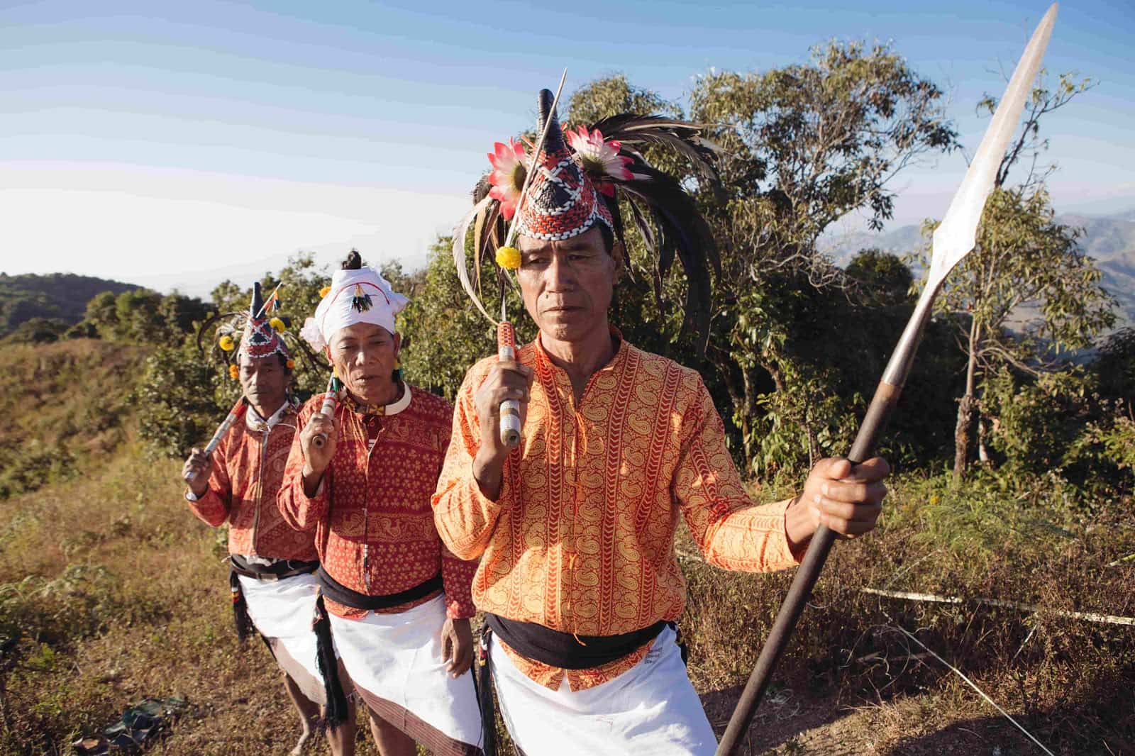 Maring men in traditional attire with feathered headgear and a spear on a hillside